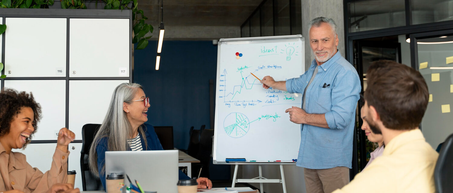 Male leader reviewing idea with engaged team Male leader standing and pointing at whiteboard while listening to a team member provide feedback, team sitting at table